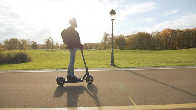 Young male moving along asphalt road with marking, passing by green lawn with evergreen and autumn trees, street lights, bench, garbage cans on sunny day. High quality 4k footage