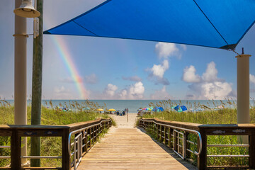 A beautiful summer landscape at the beach with a wooden footpath, people on the beach with colorful umbrellas, blue ocean water, blue sky, clouds and a rainbow in Carolina Beach North Carolina USA