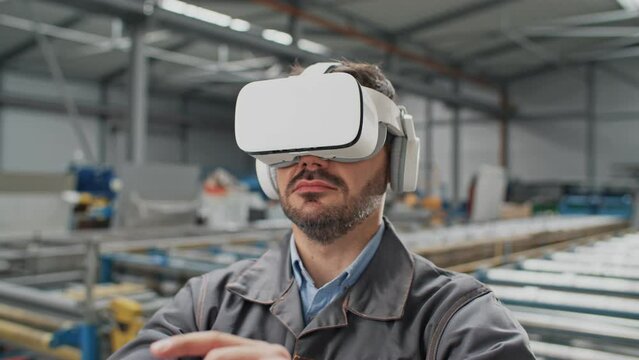 Bearded worker with uniform wearing virtual reality glasses while standing in warehouse. Busy man using technology innovation for work. Constructing new department for production in plant with headset