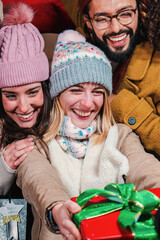 Vertical portrait of a excited woman giving a santa christmas gift to her friend. Cheerful female handing a traditional noel xmas present. Young girl holding a wrapped box to celebrate a birthday