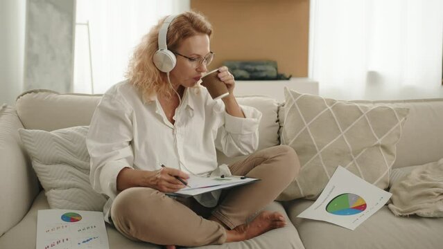Cute mature female working at home, listening to music, holding cardboard cup, sitting in yoga pose, looking attentively into papers with colorful diagrams. High quality 4k footage