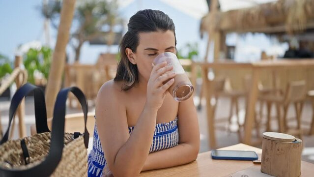 Young hispanic woman drinking iced coffee at sunny restaurant terrace