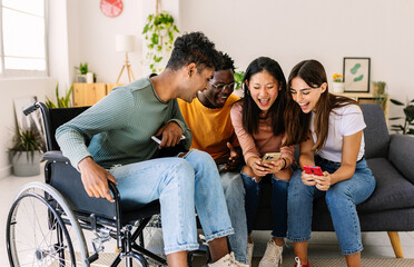 Diverse group of young friends laughing using mobile phone together at home. Inclusion concept with young indian man in wheelchair having fun with friends watching social media content on cell - obrazy, fototapety, plakaty