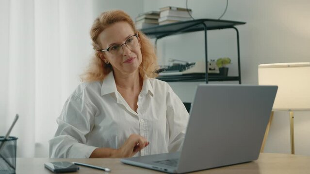 Beautiful well-groomed blonde woman in glasses finishes typing on laptop, closes lit and walks away form her workplace in a room with modern renovation. High quality 4k footage