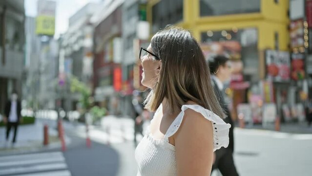 Beautiful hispanic woman in glasses, casually standing and happily smiling, posing her confidence on tokyo's modern streets, looking around, enjoying japan's urban cityscape.