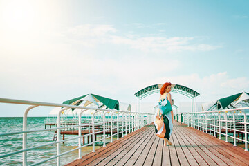 A picturesque scene unfolds as a red-haired woman, dressed in a summer dress and carrying a backpack, ambles along the resort pier with the sea providing a stunning backdrop.
