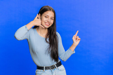 Fototapeta na wymiar Call me, here is contact number. Caucasian young woman looking at camera doing phone gesture asking for conversation. Hotline online service proposition advertisement. Girl isolated on blue background