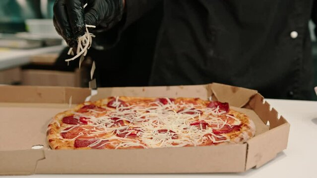 close-up A male pizza chef sprinkles mozzarella cheese on a pizza lying in a delivery box in the kitchen of a pizzeria 