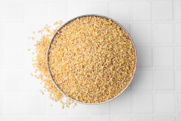 Raw bulgur in bowl on white tiled table, top view