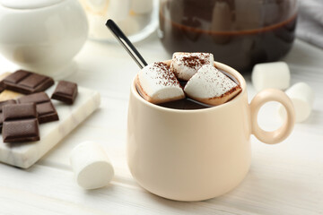 Cup of aromatic hot chocolate with marshmallows and cocoa powder on white wooden table, closeup