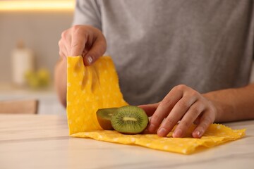 Man packing fresh kiwi into beeswax food wrap at light marble table in kitchen, closeup