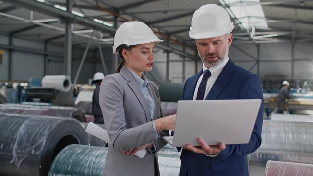 Hardworking Caucasian people with stylish suits wearing safety helmets while working in manufacturing warehouse. People using laptop while talking with each other. Partnership. Industrial factory.
