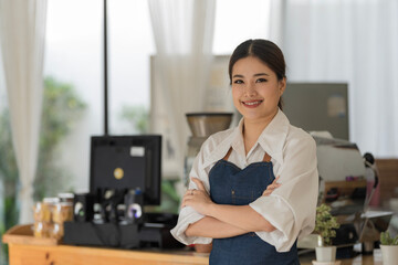Asian female barista wearing a jean apron crossed her arms at the counter bar with a smiling face,...