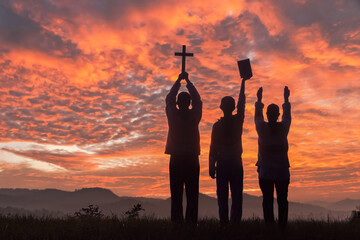 Silhouette people holding christian cross and bible for worshipping God on the mountain sunset sky background.