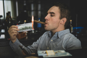 Young handsome man lighting his cigar with 100 dollar note against background of bottles in the bar. russian mafia - obrazy, fototapety, plakaty