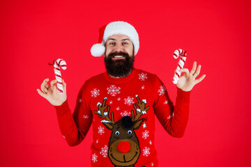 Winter holiday for man in Santa hat. Xmas sweater. Man with candy cane isolated on red. Merry Christmas. Happy New Year. Xmas holiday celebration. Santa Claus man at New Year eve. Jolly St Nicholas