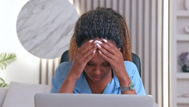 Young puzzled tired African American business woman is stressed because of mistake made or incorrectly filled out tax return and purses lips touching face with palms sits at desk in home office.