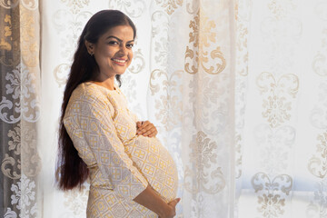 Happy pregnant indian woman touching big belly near window at home. people and expectation concept