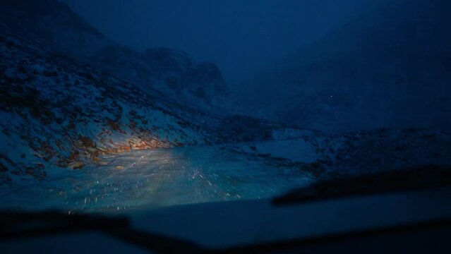 Scary road during night with heavy snowfall in mountains. POV shot of car driving on dangerous road. Winter road extreme conditions. Slippery snowy highway at night. Winter in mountains.