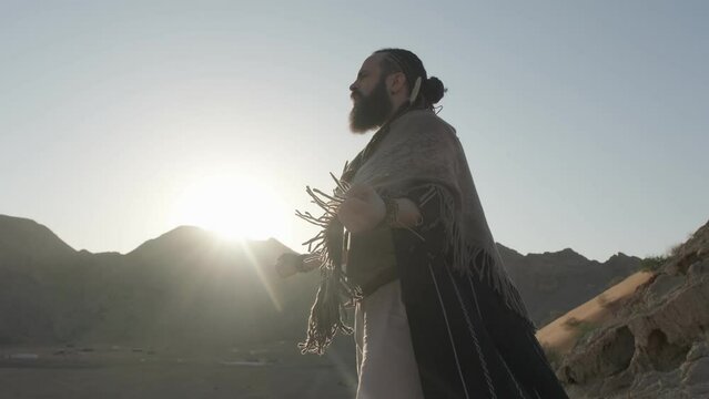A young bearded shaman prays against the backdrop of sunset among the sand dunes of the desert