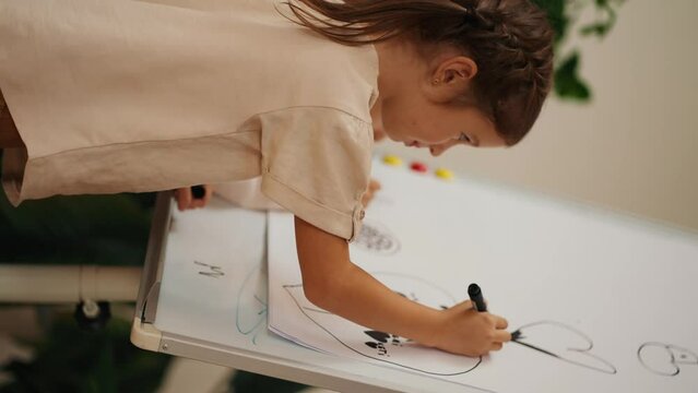 Vertical video of a girl in a white T-shirt drawing with a black marker on a large sheet of paper that is installed on a magnetic board in the room of the club for preparing preschoolers for school