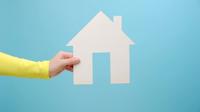 Closeup of woman hand holding white paper house, concept of dream home purchase, mortgage, real estate insurance, repairing service concept, posing isolated over blue color background wall in studio