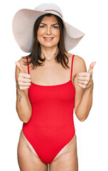 Beautiful brunette woman wearing swimsuit and summer hat success sign doing positive gesture with...