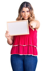 Young caucasian woman holding empty white chalkboard pointing with finger to the camera and to you, confident gesture looking serious