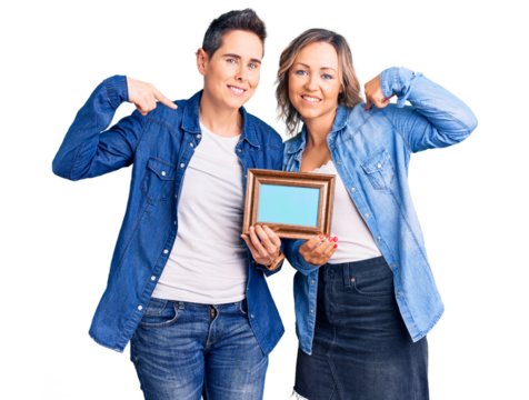 Couple of women holding empty frame looking confident with smile on face, pointing oneself with fingers proud and happy.