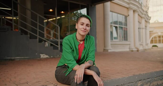 Portrait of a lesbian girl with short green hair, tattoos, in a Green shirt and black pants who sits on a small step on the street near the building
