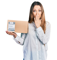 Beautiful caucasian woman holding delivery package covering mouth with hand, shocked and afraid for...