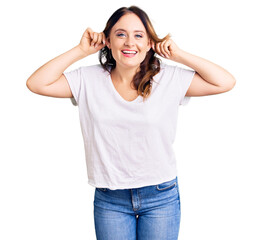 Young beautiful caucasian woman wearing casual white tshirt smiling pulling ears with fingers,...