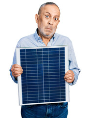Senior man with grey hair and beard holding photovoltaic solar panel clueless and confused...