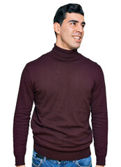 Fototapeta premium Handsome hispanic man wearing casual turtleneck sweater looking away to side with smile on face, natural expression. laughing confident.