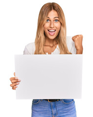 Fototapeta premium Beautiful blonde young woman holding blank empty banner screaming proud, celebrating victory and success very excited with raised arms