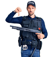 Young handsome man wearing police uniform holding shotgun with angry face, negative sign showing dislike with thumbs down, rejection concept