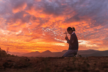 Human kneeling down praying and holding christian cross for worshipping God at sunset sky...