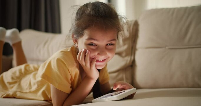 Close-up shot: Portrait of a happy little brunette girl lying on a light brown sofa looking at something on her phone smiling and looking at the camera in a modern room