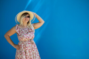 A pretty smiling woman in summer against a rich blue wall in glasses and a sundress Copy space. A beautiful woman stands against a blue wall with sun rays Summer concept advertising flyer copy space.