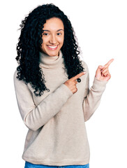 Young hispanic woman with curly hair wearing turtleneck sweater smiling and looking at the camera pointing with two hands and fingers to the side.