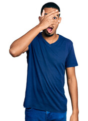 Young african american man wearing casual t shirt peeking in shock covering face and eyes with hand, looking through fingers with embarrassed expression.