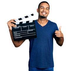 Young african american man holding video film clapboard smiling happy and positive, thumb up doing...