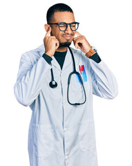 Young african american man wearing doctor uniform and stethoscope covering ears with fingers with annoyed expression for the noise of loud music. deaf concept.