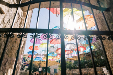 View through a wrought-iron fence to colorful umbrellas hanging above the street of an ancient town