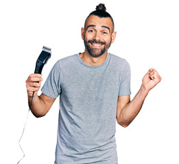 Hispanic man with ponytail holding electric razor machine screaming proud, celebrating victory and...