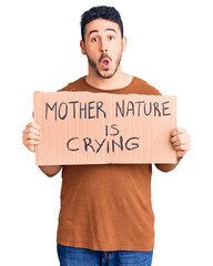Young hispanic man holding mother nature is crying protest cardboard banner scared and amazed with open mouth for surprise, disbelief face