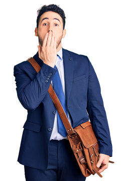 Young hispanic man wearing suit and leather bag covering mouth with hand, shocked and afraid for mistake. surprised expression