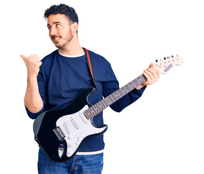 Young hispanic man playing electric guitar pointing thumb up to the side smiling happy with open mouth