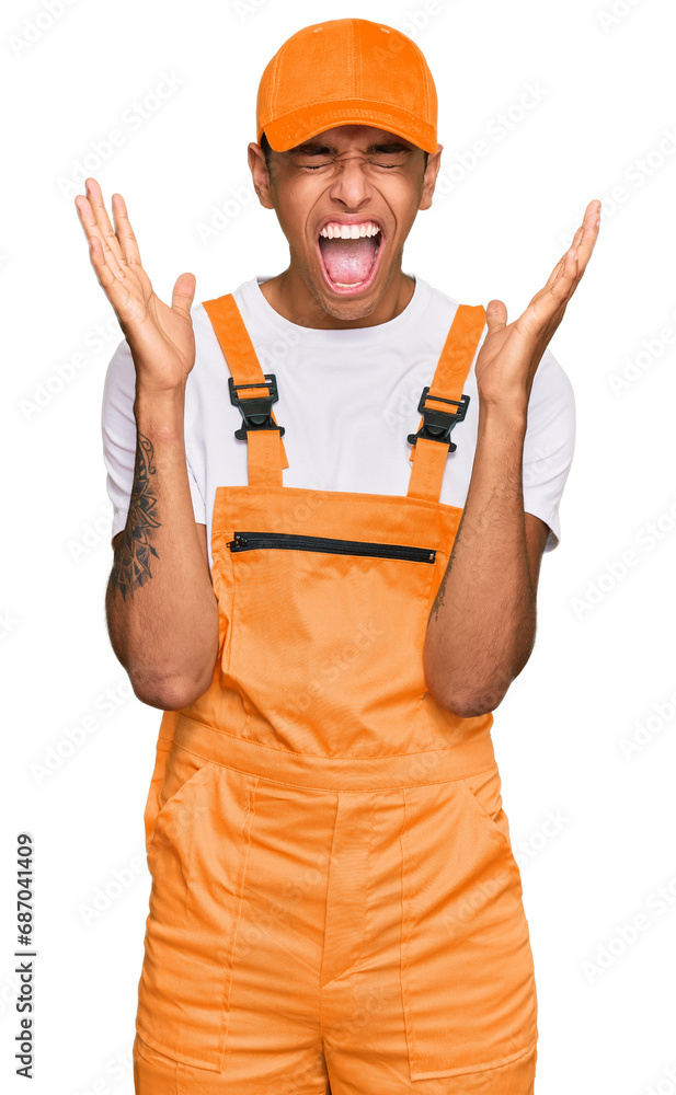 Wall mural Young handsome african american man wearing handyman uniform celebrating mad and crazy for success with arms raised and closed eyes screaming excited. winner concept