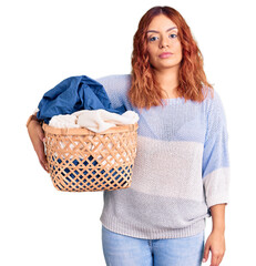 Young latin woman holding laundry basket thinking attitude and sober expression looking self confident